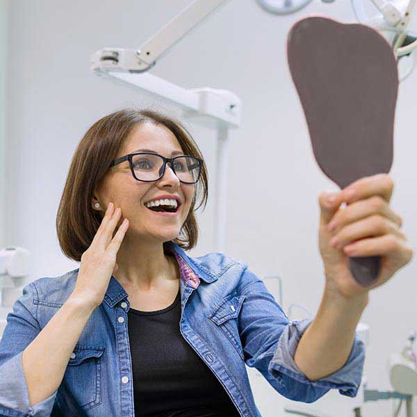 a woman wearing glasses looks into a small mirror with excitement after a dental visit