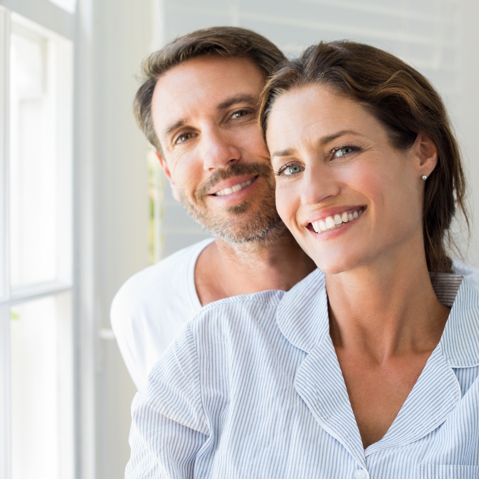 Happy young couple sitting near window in the bedroom. Portrait of smiling man and woman looking at camera at home. Couple in pajamas during the morning.