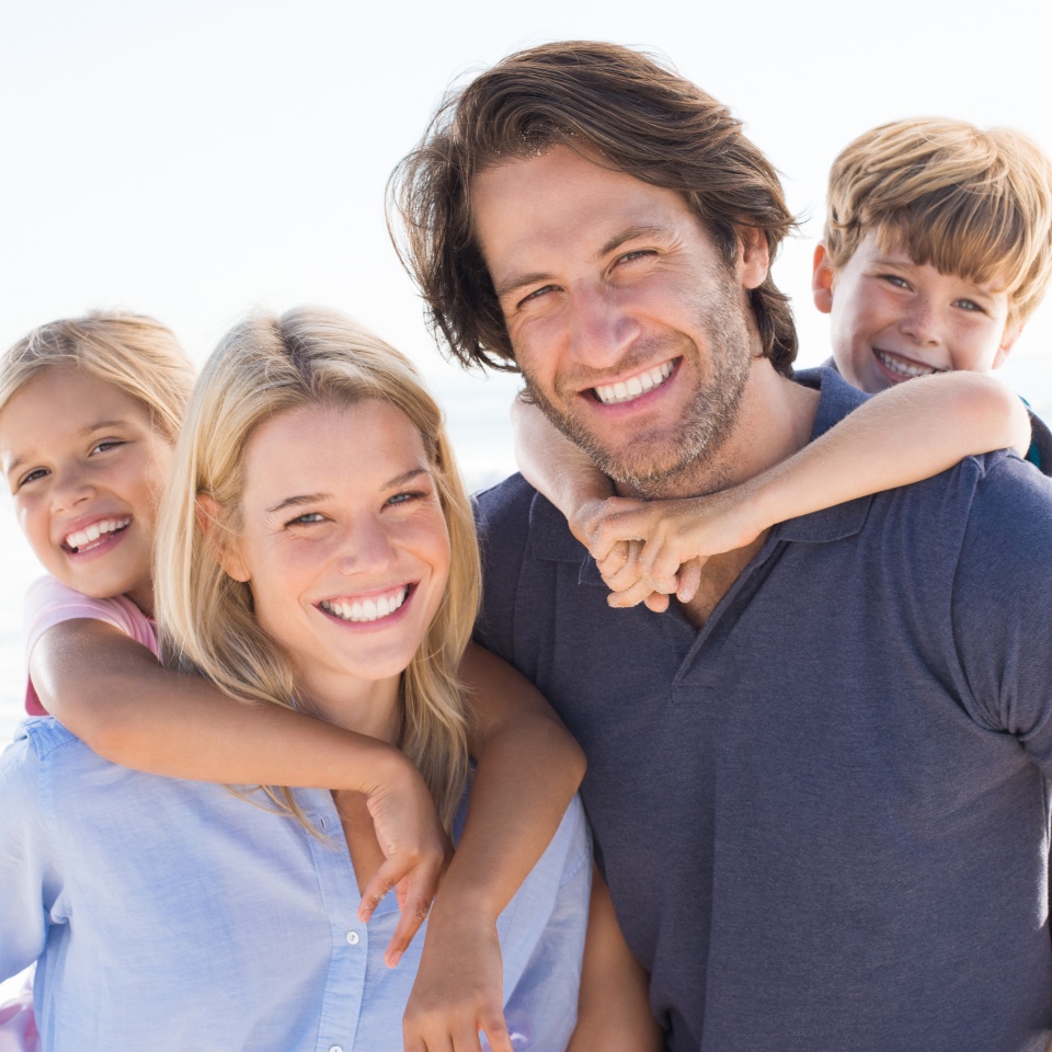 Parents giving piggyback ride to kids at beach. Close up of smiling family having fun at summer vacation. Portrait of happy family looking at camera at beach.
