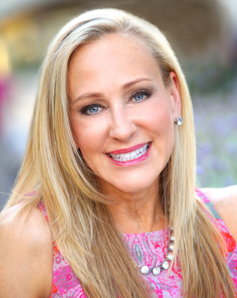 A woman with long blonde hair and blue eyes is smiling at the camera. She is wearing a pink patterned top, a necklace, and stud earrings. The background is softly blurred.