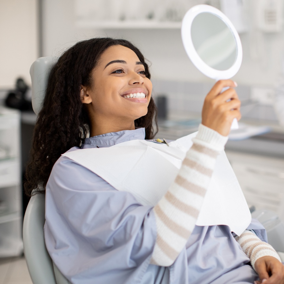 Happy Black Female Patient Looking At Mirror After Dental Treatment In Clinic, Cheerful African American Woman Sitting In Chair In Stomatological Cabinet And Enjoying Her Beautiful Smile, Closeup