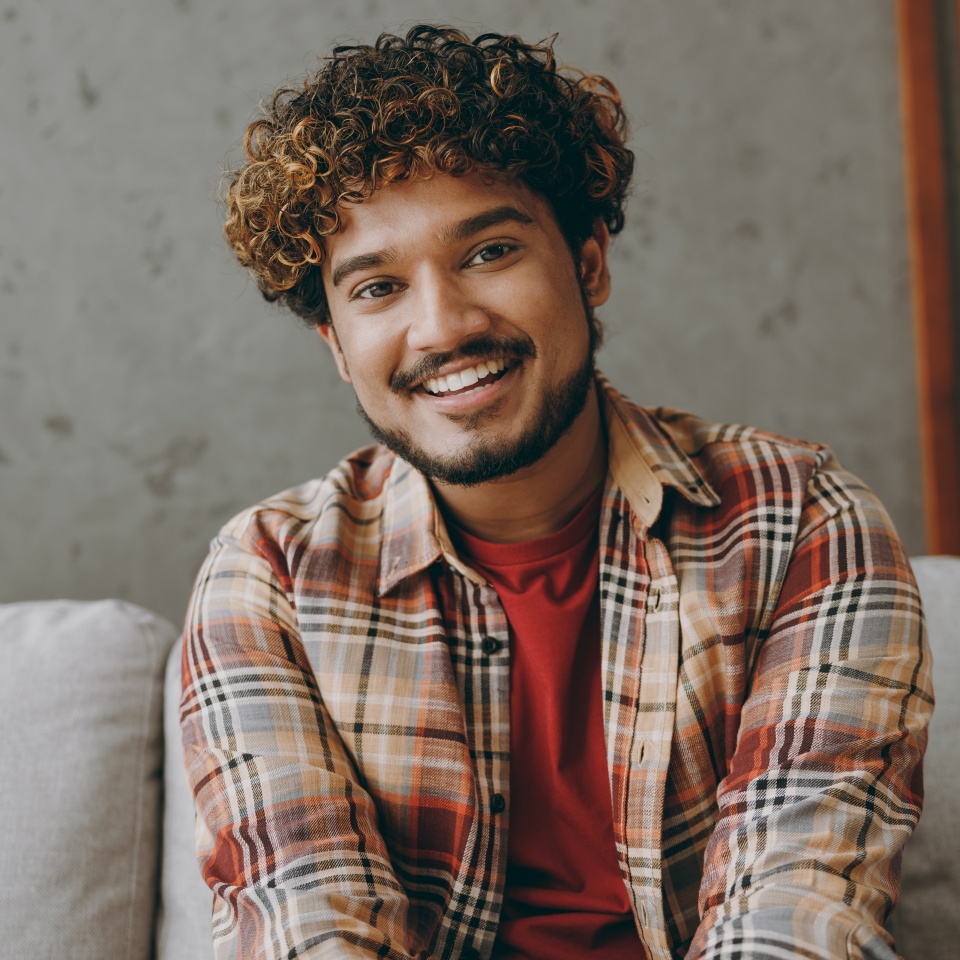 Close up young smiling happy Indian man wears casual clothes look camera sits on grey sofa couch stay at home hotel flat rest relax spend free spare time in living room indoor. People lounge concept