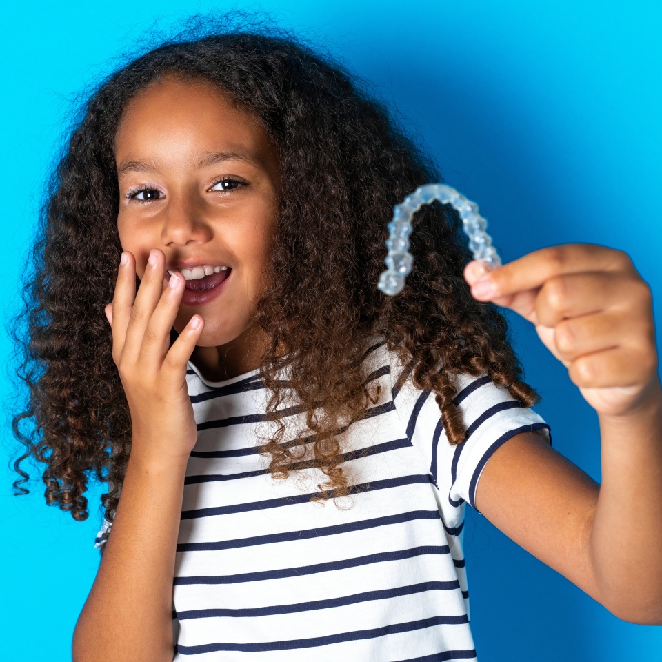 Happy beautiful kid girl with afro curly hairstyle wearing striped T-shirt holding and showing at camera an invisible aligner while laughing. Dental healthcare and confidence concept.