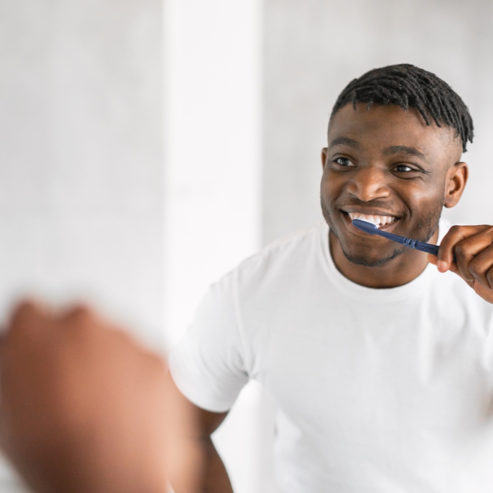 Handsome African American millennial guy brushing teeth with toothbrush stands by the mirror, caring for his oral health in modern bathroom. Daily dental care routine. Selective focus