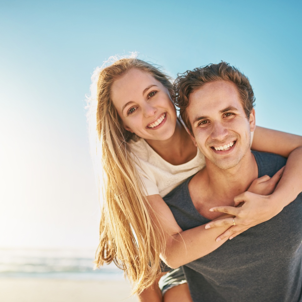 Portrait, piggy back and beach with couple, smile and sunshine with happiness, honeymoon and romance. Face, seaside and man carrying woman with vacation, journey and summer with getaway trip or love