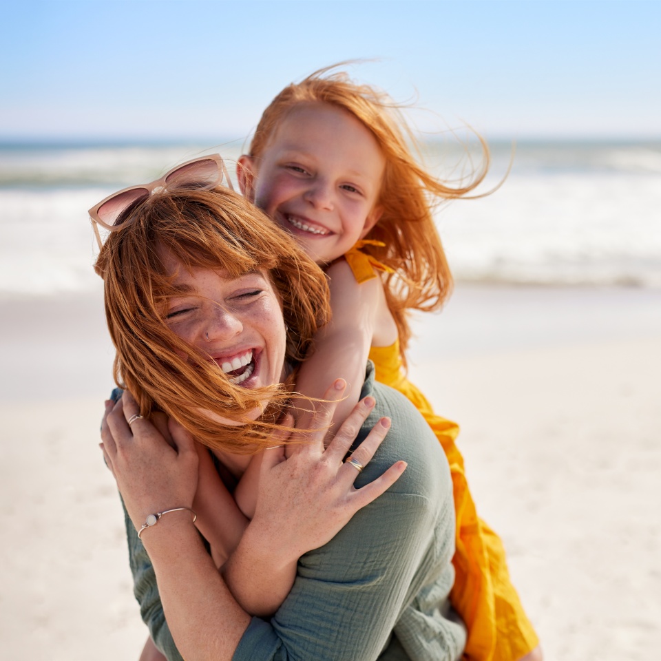 Smiling mother and beautiful daughter having fun at the beach. Portrait of happy woman giving a piggyback ride to cute little girl with copy space. Portrait of happy red hair kid embracing her mom.