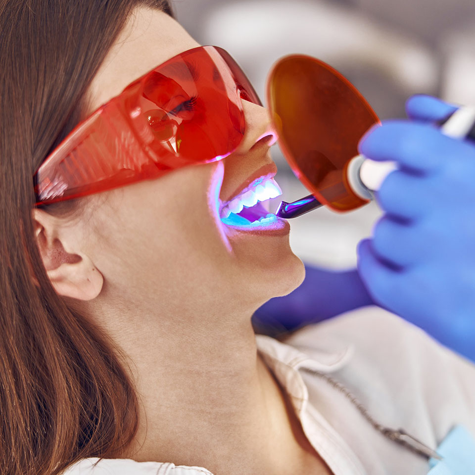Woman wearing red protective glasses receives a dental treatment with a curing light, administered by a gloved dental professional.