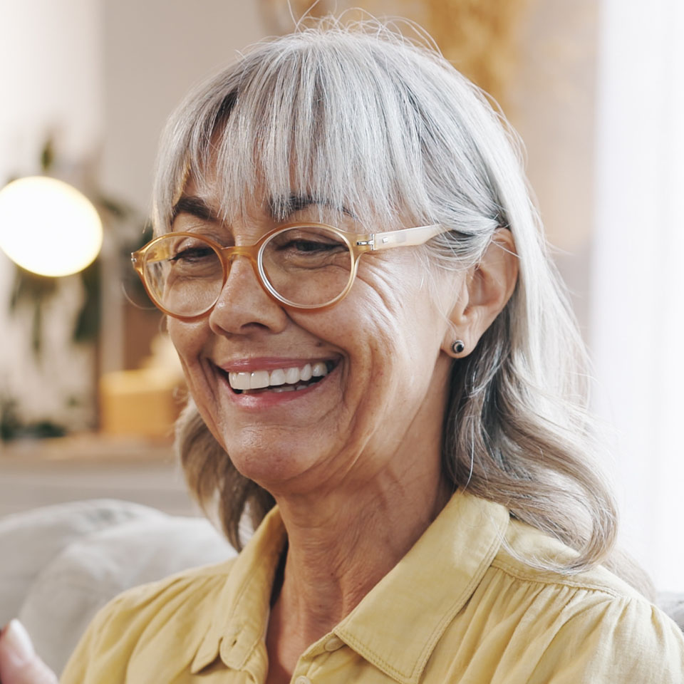 Older woman with gray hair and glasses, wearing a yellow shirt, smiling indoors with a softly lit background.