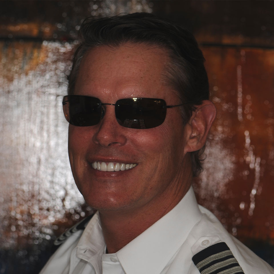 Man wearing a white pilot uniform and dark sunglasses, smiling in front of a textured, brown-toned background.