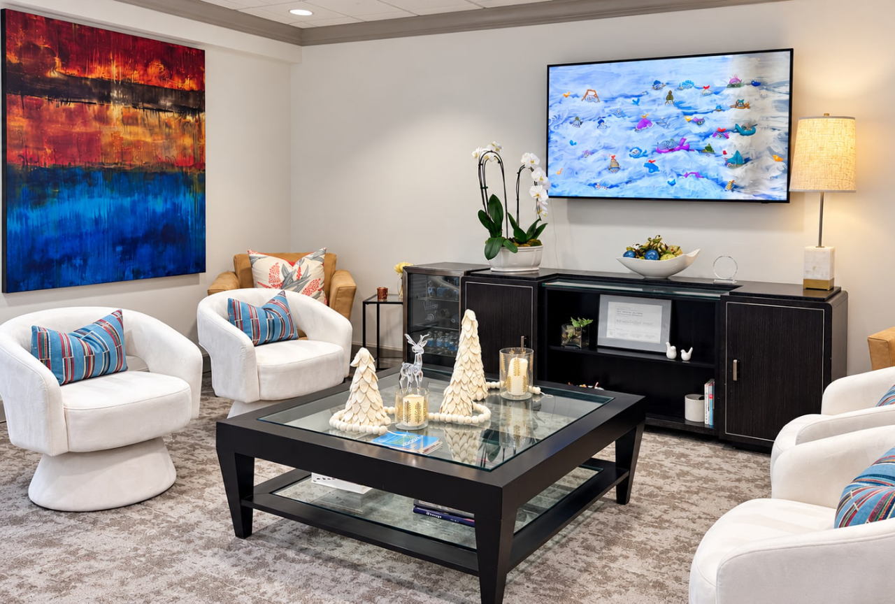 A waiting area with white armchairs, a black coffee table decorated with seasonal ornaments, a television displaying a winter scene, and a large abstract painting on the wall.